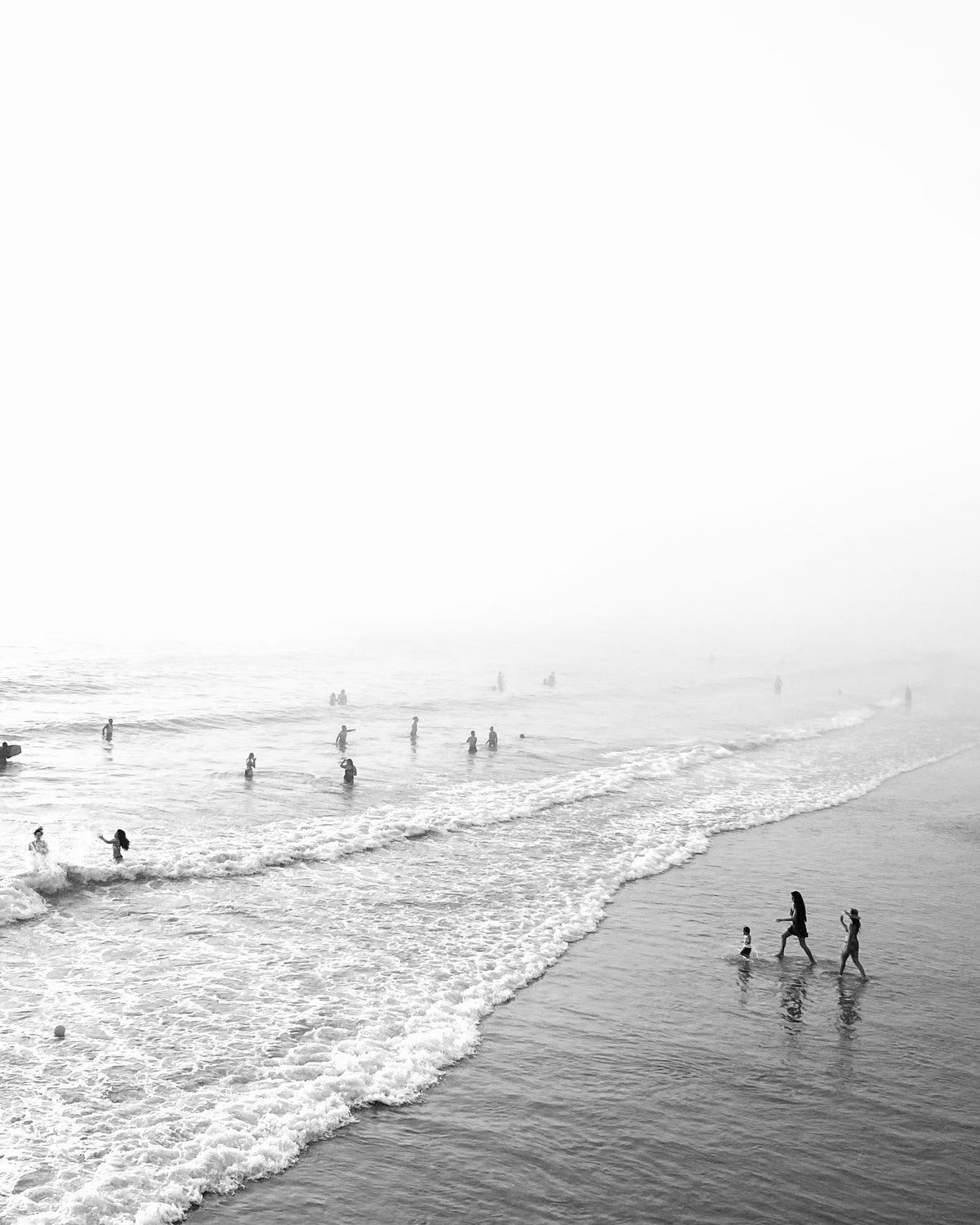 A family wading into the surf on a misty beach, with swimmers in the waves, in black and white